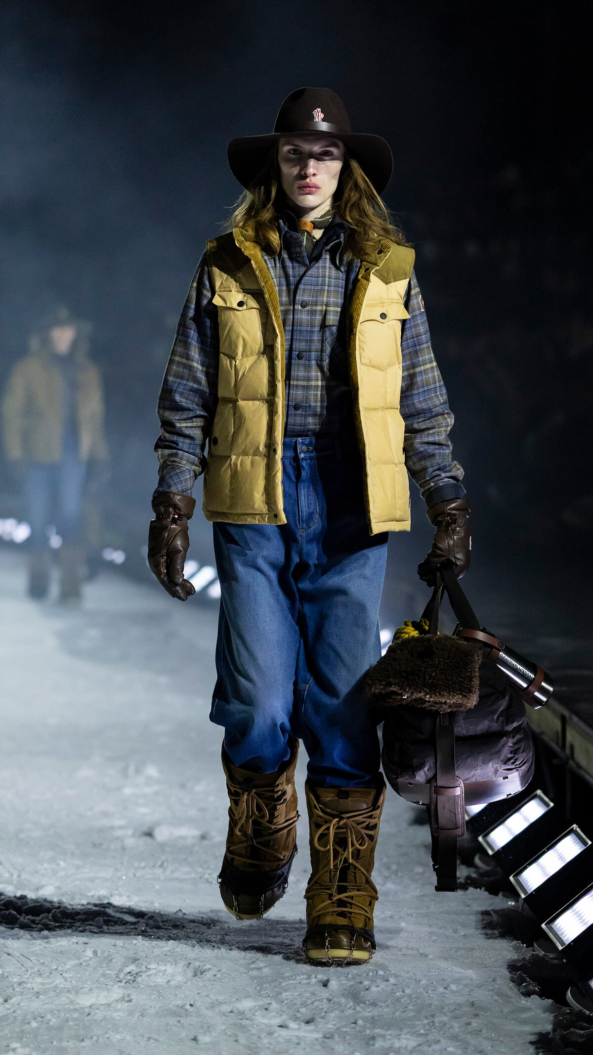 Model walking on an icy runway at night in a tan puffer vest, plaid shirt, blue jeans, fur-lined boots, and a wide-brim hat, carrying a duffel bag.
