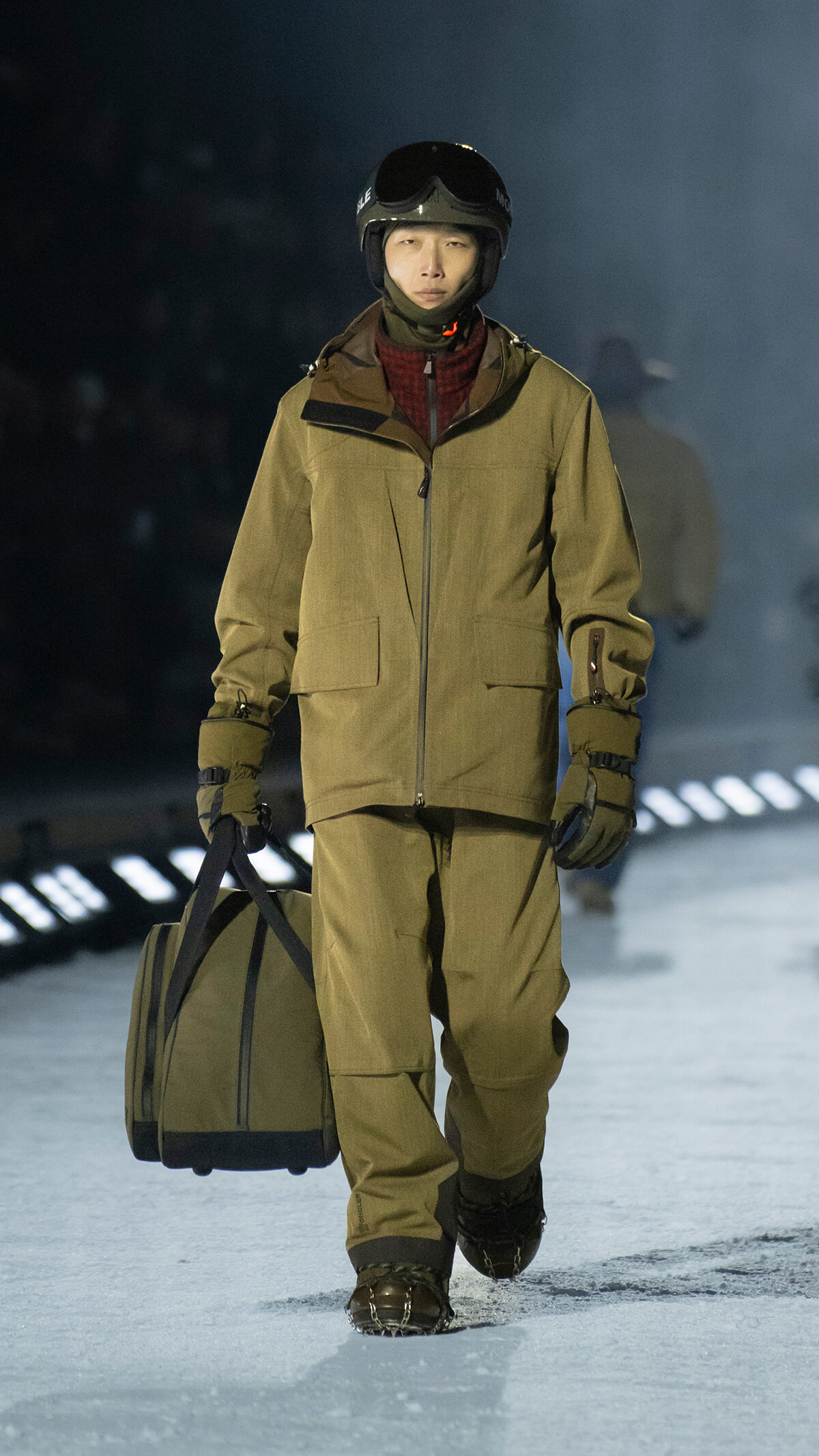Person in a tan winter jacket with helmet and goggles walks on snow, carrying a large duffel bag on their side.