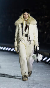 Model walking on a snowy runway in a cream quilted snowsuit with a fur collar, holding a white ski helmet.**I will fix**