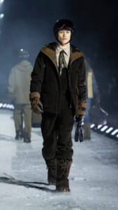 Model walking on a snowy runway in a dark winter outfit with helmet, goggles, scarf, and brown gloves.