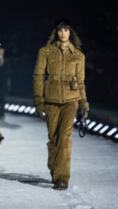 Female model walking on a snowy runway in a tan corduroy snowsuit with goggles, belt, and gloves.