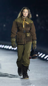 Model walking on a snow runway at night in a brown fringed Western-style jacket with floral embroidery, belt, gloves, and holding a helmet.