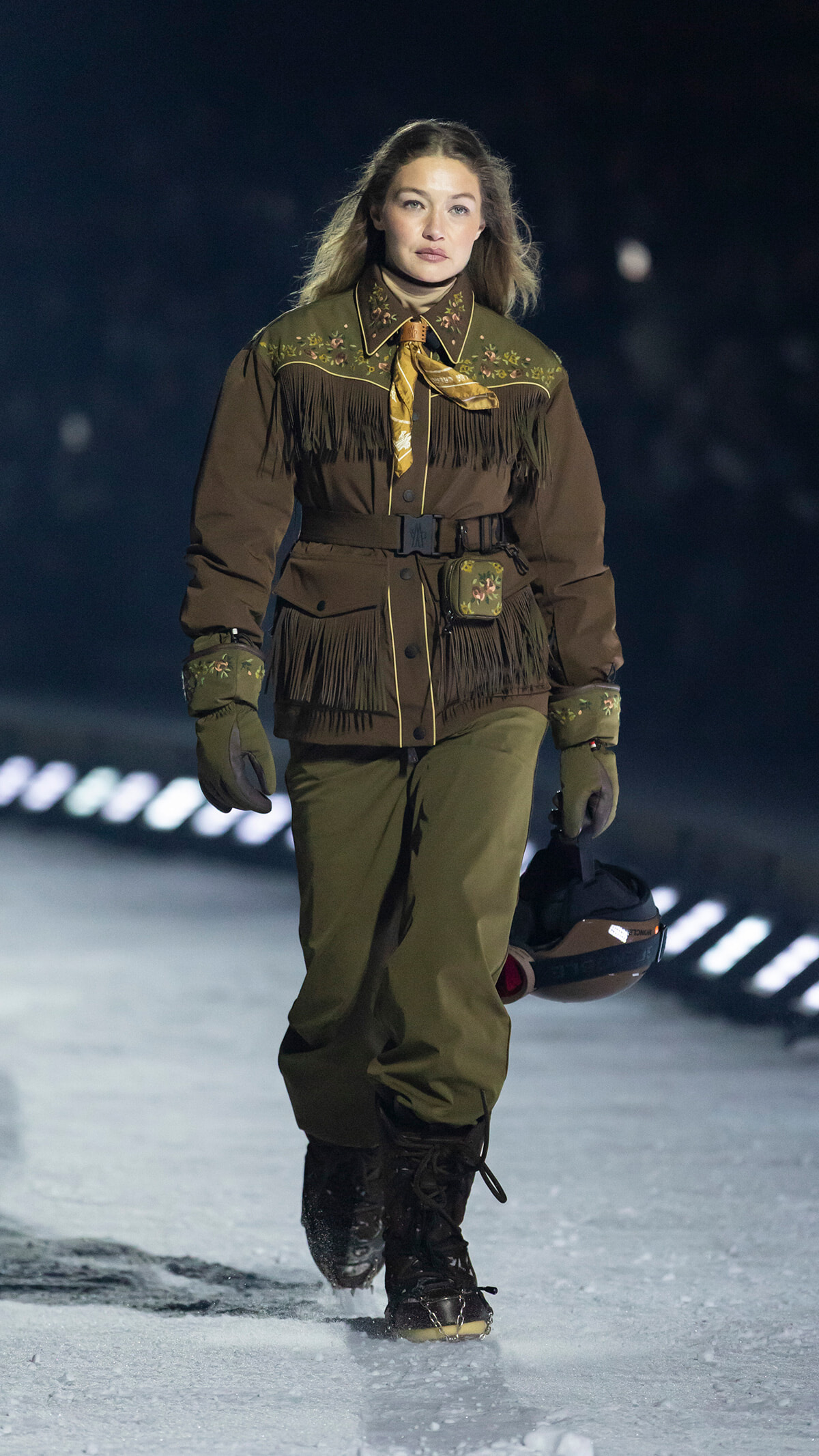 Model walking on a snow runway at night in a brown fringed Western-style jacket with floral embroidery, belt, gloves, and holding a helmet.