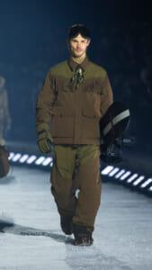 Male model walking on a snowy runway in a brown and olive fringed jacket, goggles on his head, carrying a snowboard.