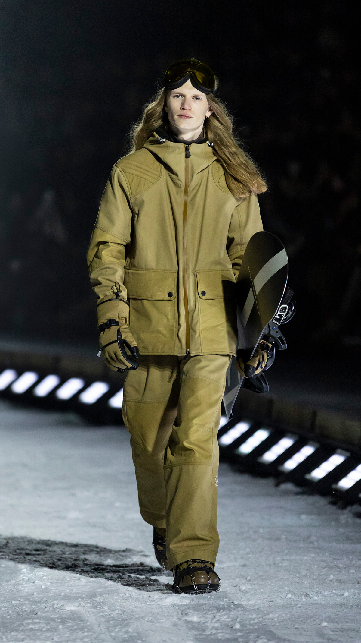 Person in tan snowboarding outfit walks on snow, holding a snowboard and wearing goggles on their head during a show/scene.