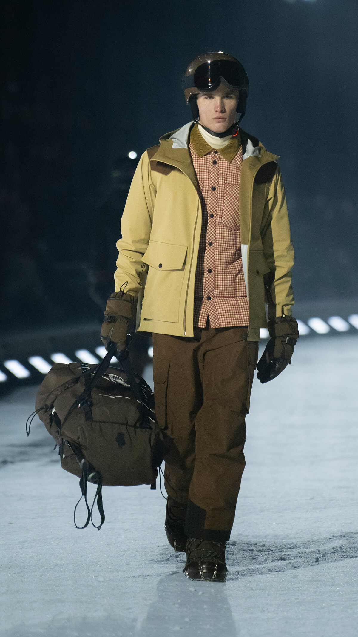 Athlete in a tan jacket and helmet walks on an ice rink, pulling a large brown duffel bag.