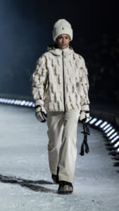 Model walking on a snowy runway at night in an off-white patterned winter jacket, matching pants, a knit beanie, and gloves.