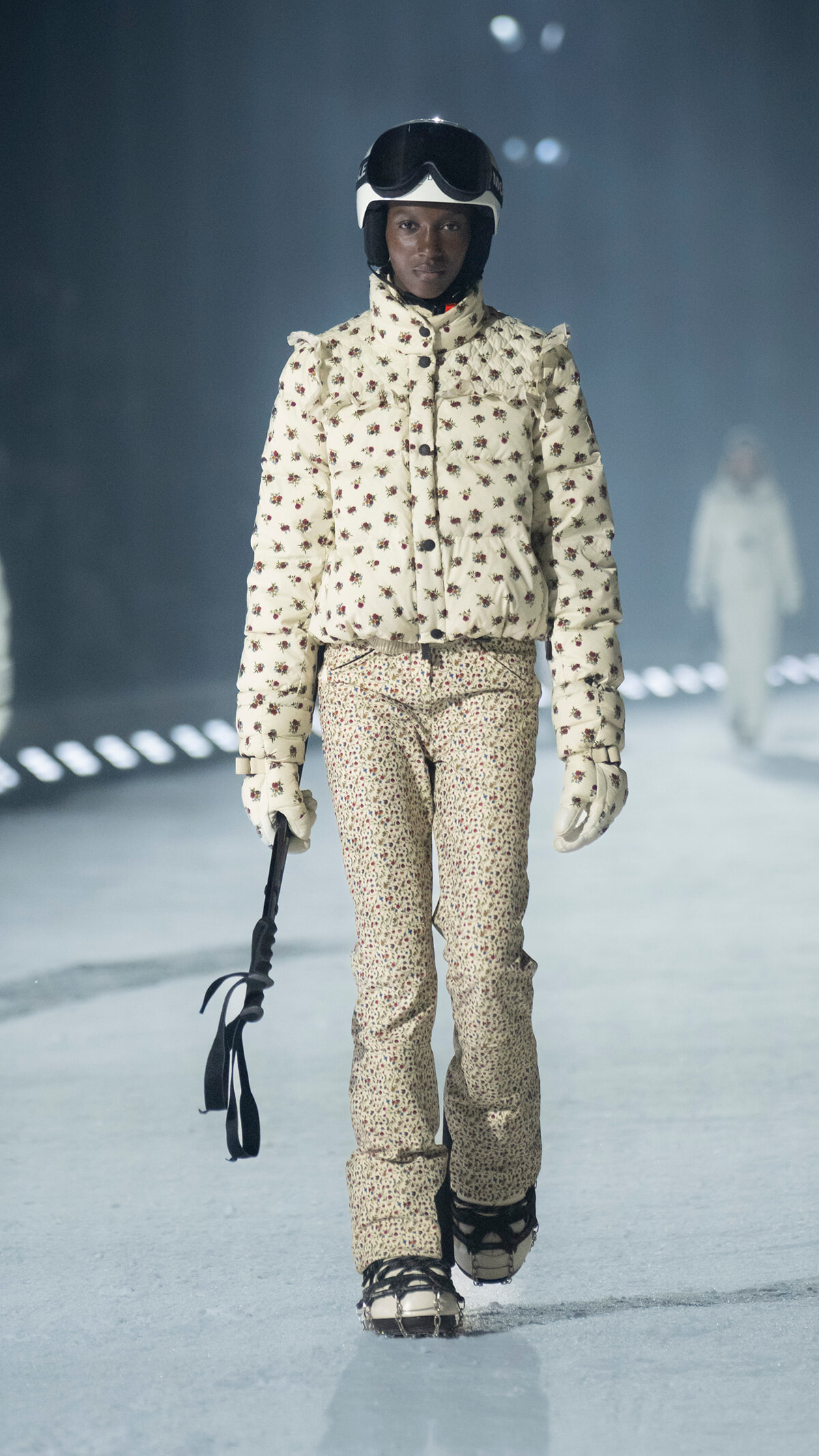 Model walking a runway in a cream floral ski suit with a helmet and goggles, holding a ski pole.