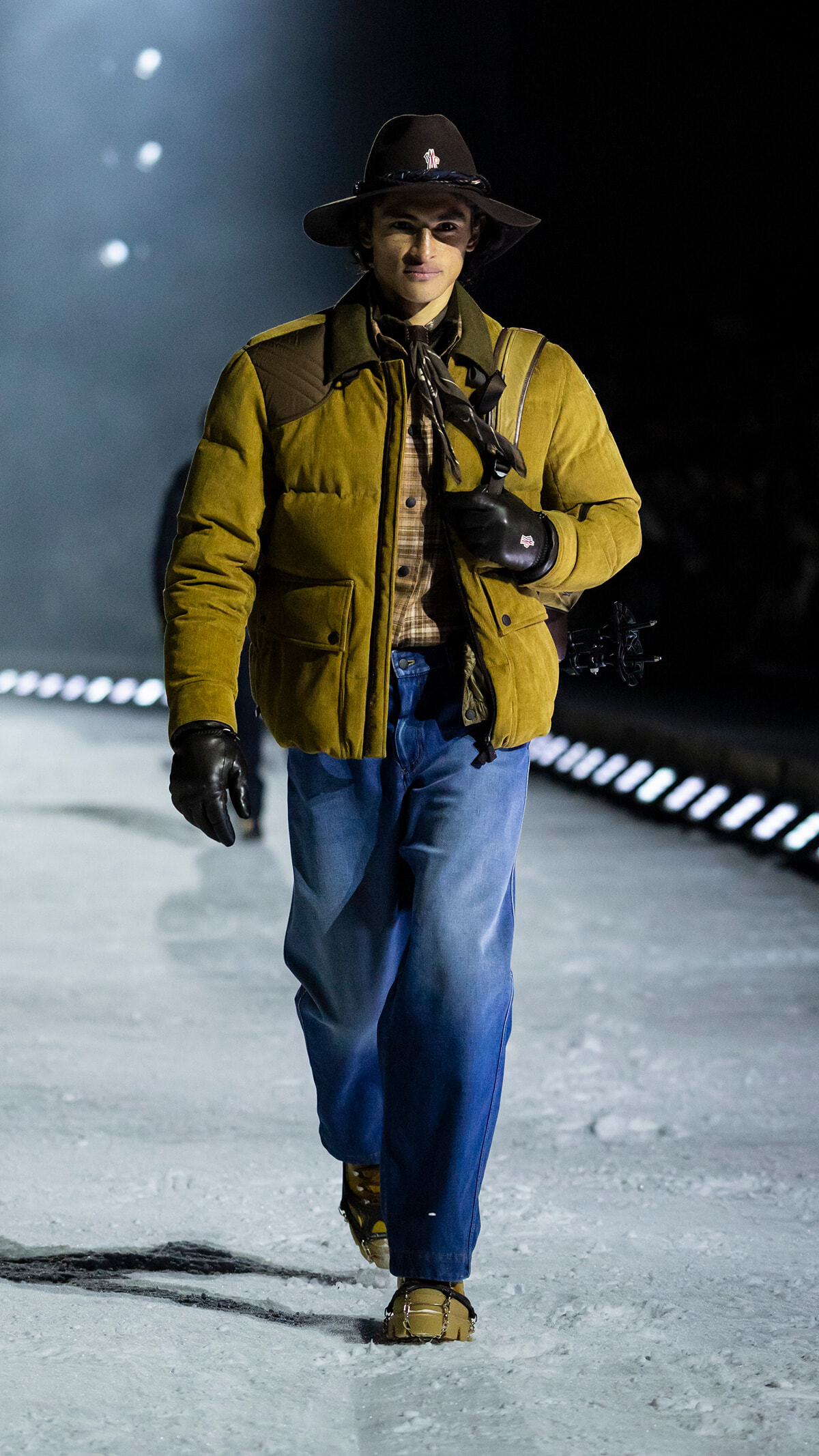 Man walking on snow at night in a tan puffer jacket, plaid shirt, jeans, gloves, and a wide-brimmed hat, carrying a bow over his shoulder.