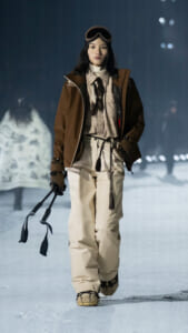 Female model walking on a snowy runway in a brown ski jacket, beige pants, and winter goggles atop her head.