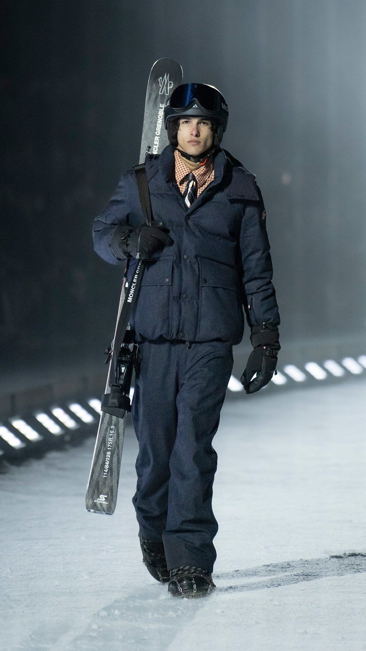 Man in a navy winter suit with helmet and goggles carrying a snowboard over his shoulder, walking on snow.
