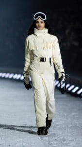 Model walking on a snowy runway in a cream fringe-parka snowsuit with a belt and gloves, wearing ski goggles.