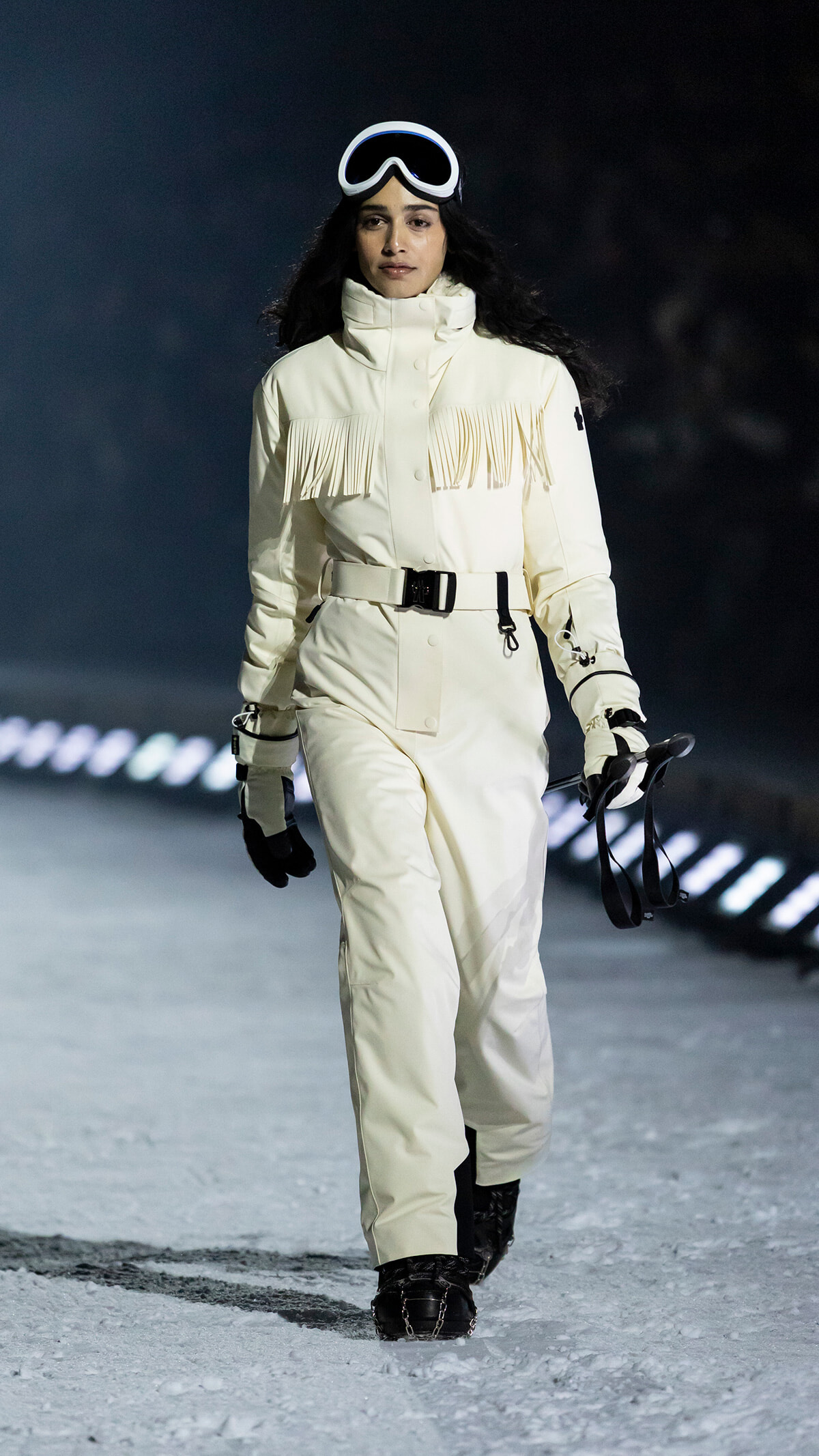 Model walking on a snowy runway in a cream fringe-parka snowsuit with a belt and gloves, wearing ski goggles.