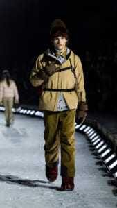 Male model walking a snowy runway at night in a tan parka, gloves, knit beanie with red ski goggles, and red boots.