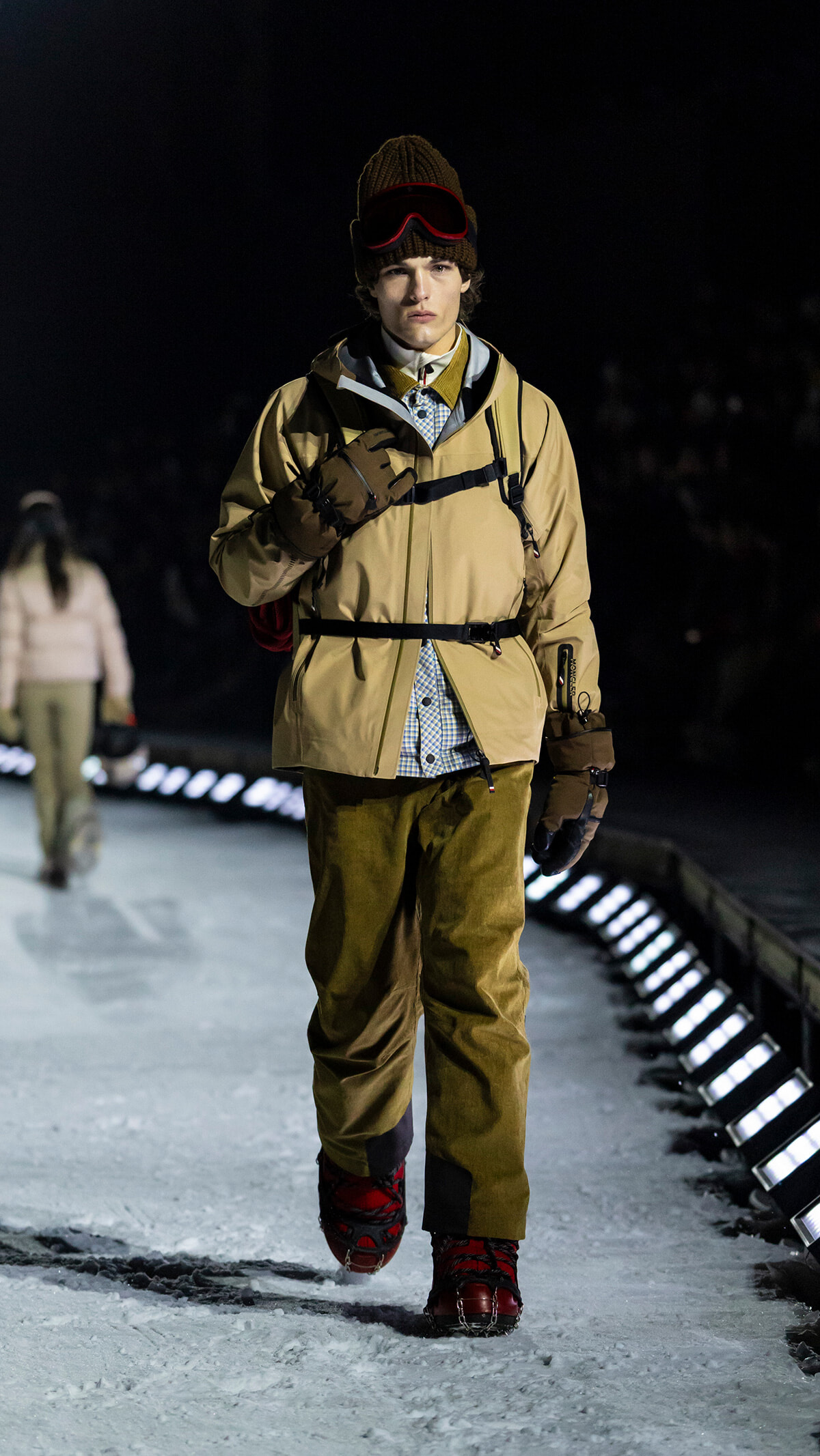 Male model walking a snowy runway at night in a tan parka, gloves, knit beanie with red ski goggles, and red boots.