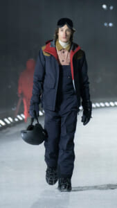 Male model in a navy ski suit with red accents, goggles on his head, carrying a black helmet while walking on an ice runway.