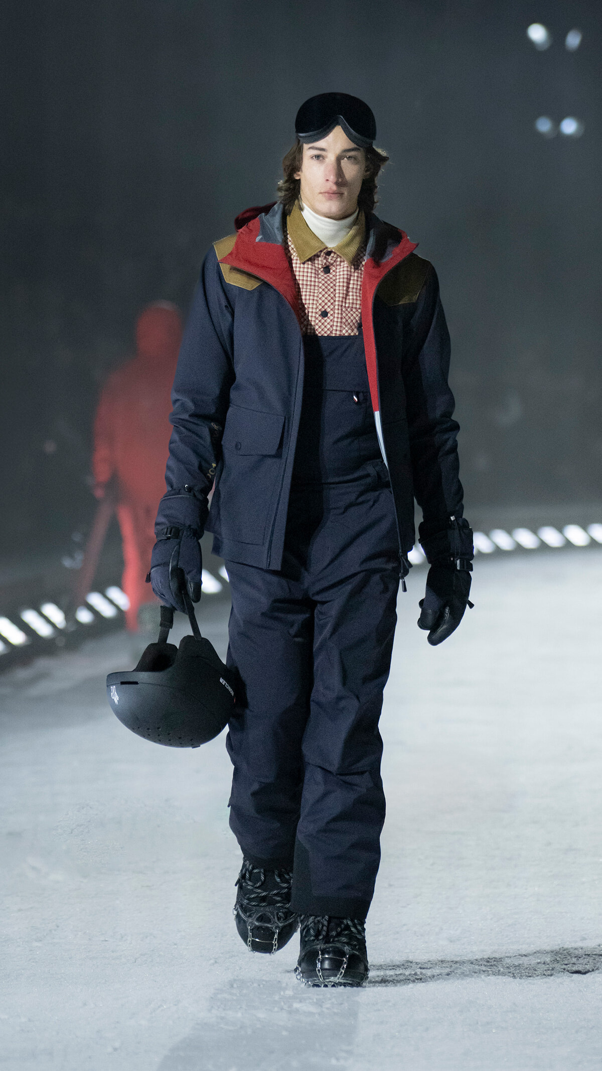Male model in a navy ski suit with red accents, goggles on his head, carrying a black helmet while walking on an ice runway.