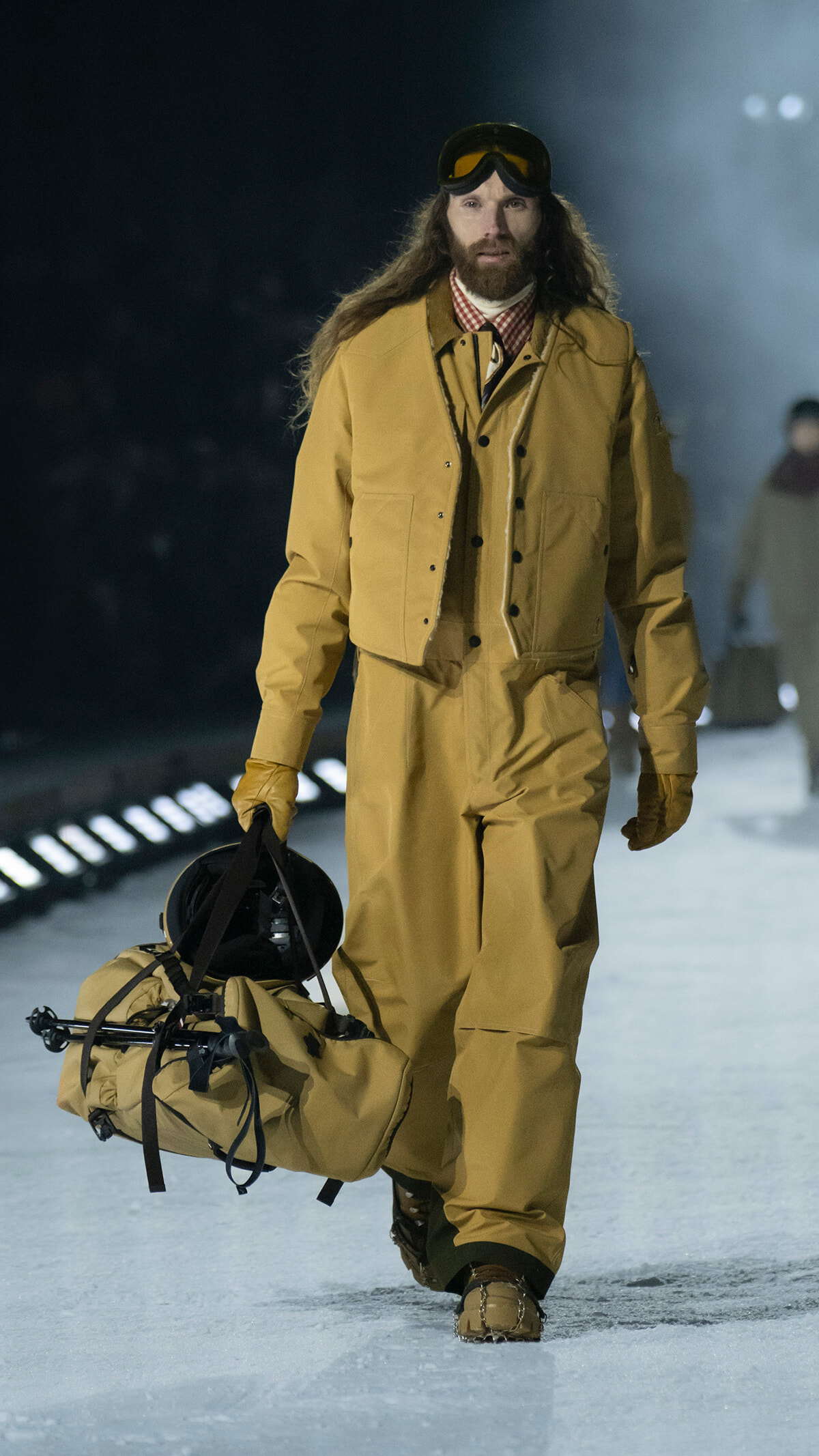 Male model with long hair and beard wearing tan snowsuit and ski goggles walks on a snowy runway, carrying a large tan duffel bag and gear.