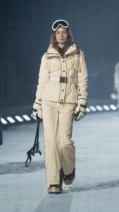 Model on a snowy runway wearing a cream fringed snowsuit, belt, gloves, and white ski goggles perched on her head.