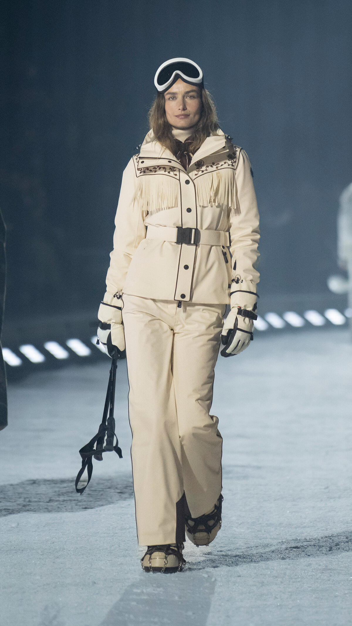Model on a snowy runway wearing a cream fringed snowsuit, belt, gloves, and white ski goggles perched on her head.