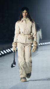 Female model walking a snowy runway in an ivory embroidered snowsuit with a belt, cream gloves, and goggles on her head, holding a ski helmet.