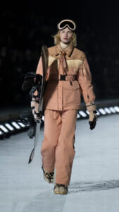 Model walking on an icy runway in a peach fringe-ski suit with goggles, carrying a snowboard and snowboard boots.