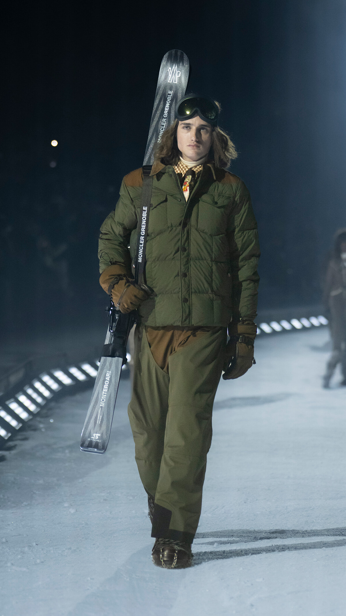 Model walks a snowy runway in a green padded jacket, brown gloves, and ski pants, carrying a pair of Moncler skis and wearing ski goggles on her head at night.