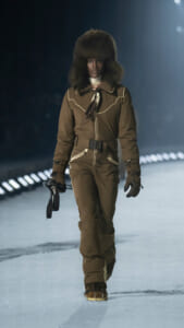 Model walking on a snowy runway in a brown, belted snowsuit with a large fur hat and gloves, holding goggles in one hand.