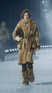 Model walking on a snowy runway in a chunky brown knit coat, scarf, mittens, and tan lace-up boots.