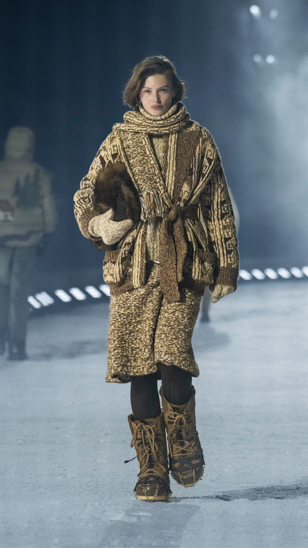 Model walking on a snowy runway in a chunky brown knit coat, scarf, mittens, and tan lace-up boots.
