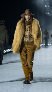 Model on a snowy runway wearing a tan fur coat, patterned knit sweater, brown pants, and a wide-brim hat.