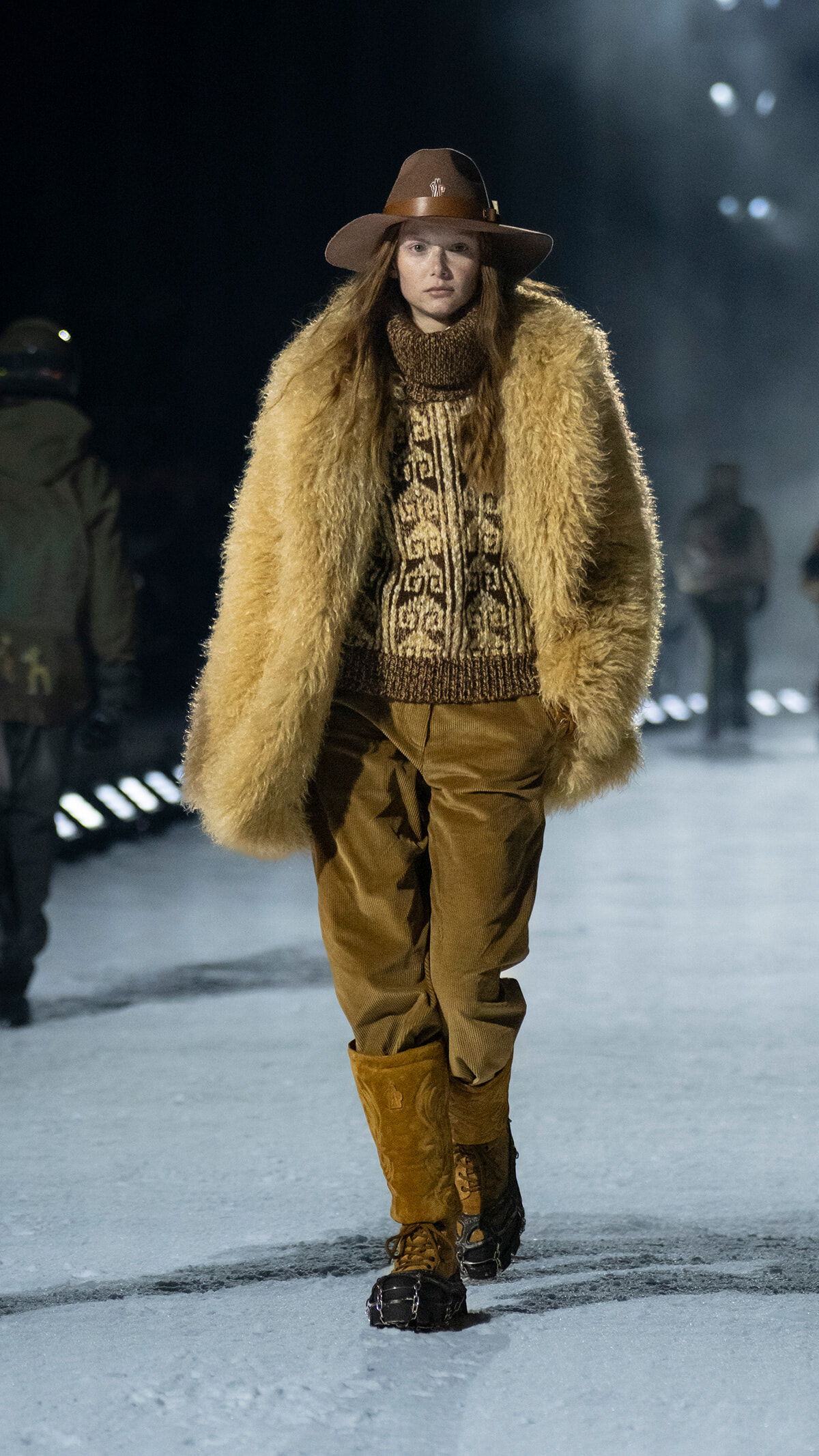 Model on a snowy runway wearing a tan fur coat, patterned knit sweater, brown pants, and a wide-brim hat.
