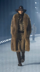 Model walking a runway in a long brown fur coat, wide-brim hat, turtleneck sweater, and tall boots.