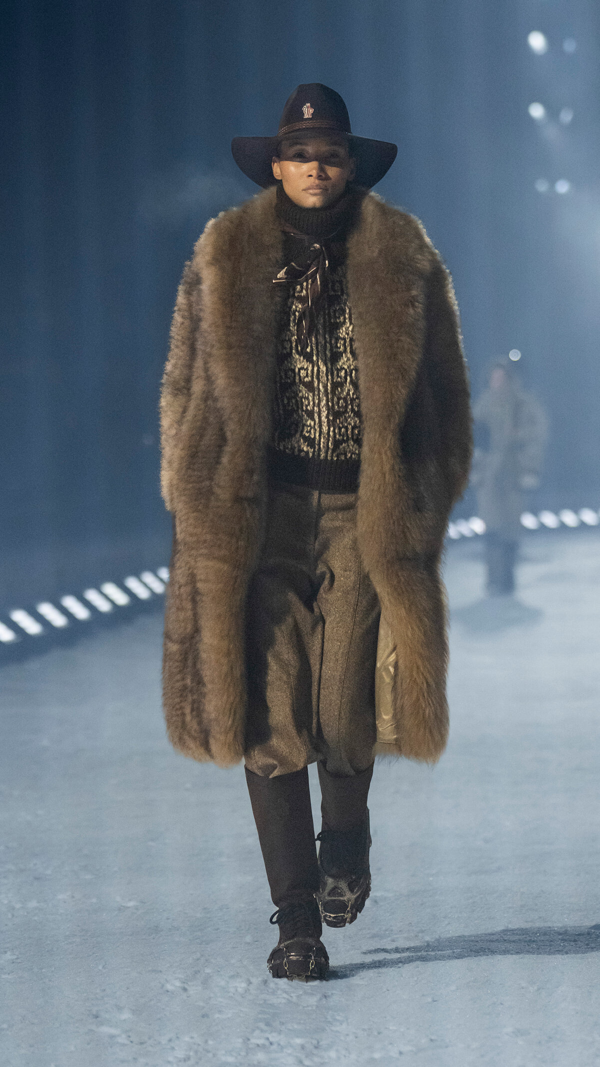 Model walking a runway in a long brown fur coat, wide-brim hat, turtleneck sweater, and tall boots.