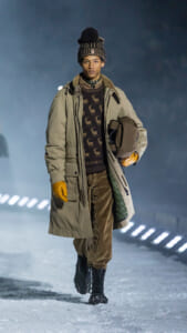 Male model walking on a snowy runway in a beige parka, brown reindeer-pattern sweater, corduroy pants, knit beanie, and orange gloves, carrying a brown helmet.