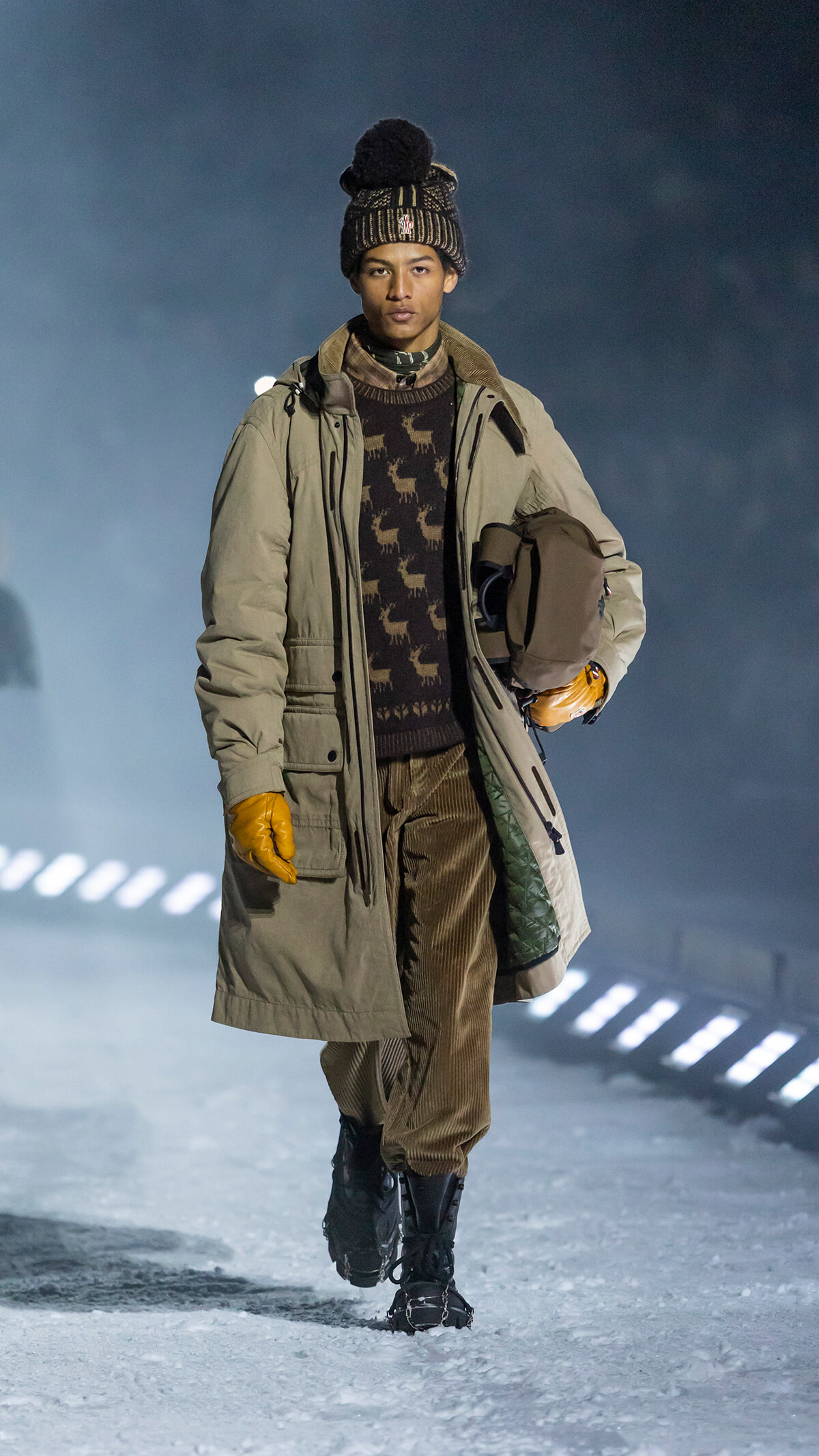 Male model walking on a snowy runway in a beige parka, brown reindeer-pattern sweater, corduroy pants, knit beanie, and orange gloves, carrying a brown helmet.