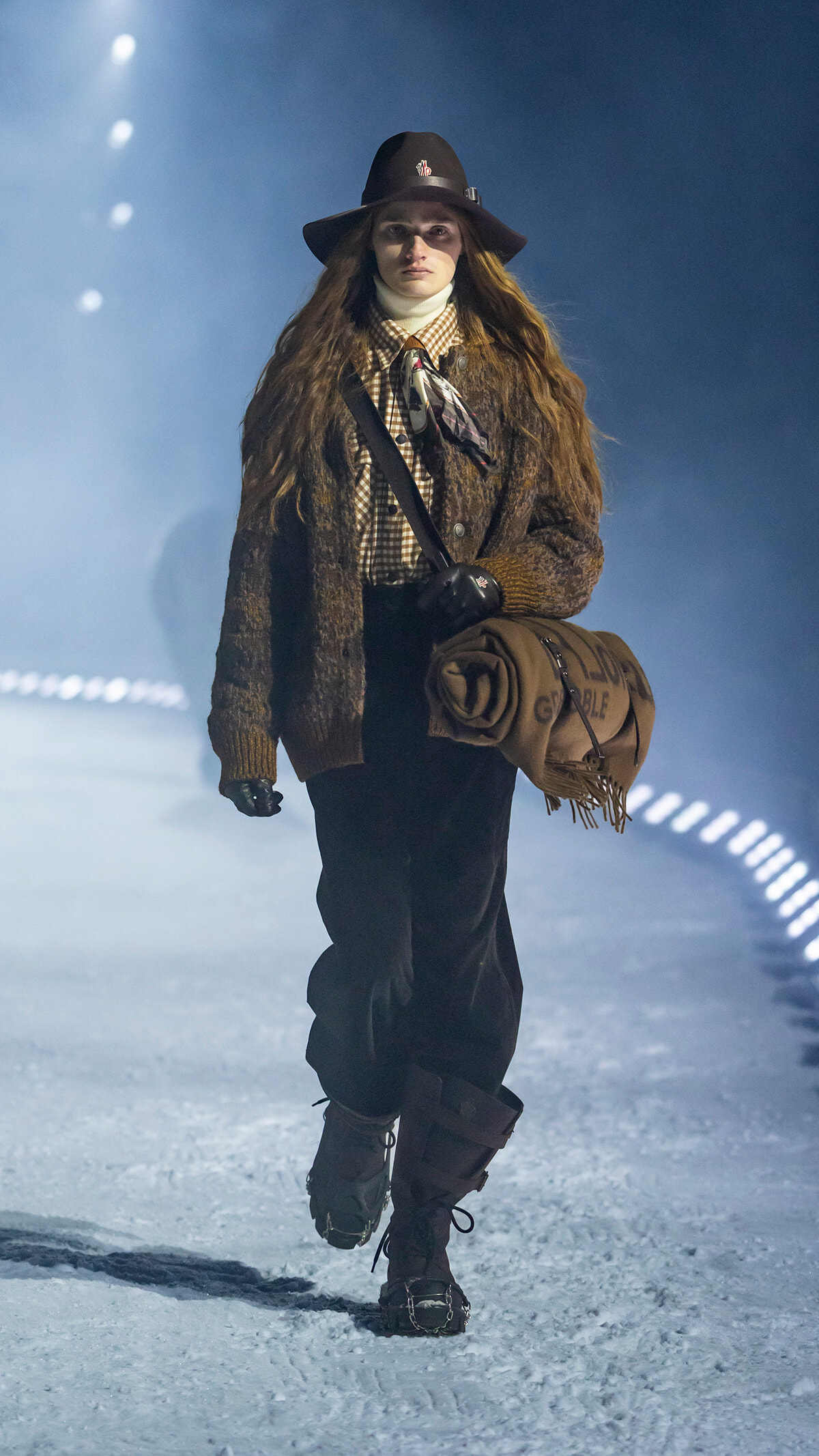 Female model walking a snowy runway in a brown fur coat, checkered shirt, scarf, and tall boots, carrying a rolled blanket under her arm.
