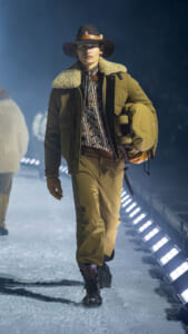 Model walking a snowy runway in an olive shearling-lined parka, brown pants, wide-brim hat, and carrying a large olive bag.