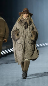 Model walking a snowy runway in a speckled oversized coat with fur collar and cuffs, brown hat, and knee-high boots