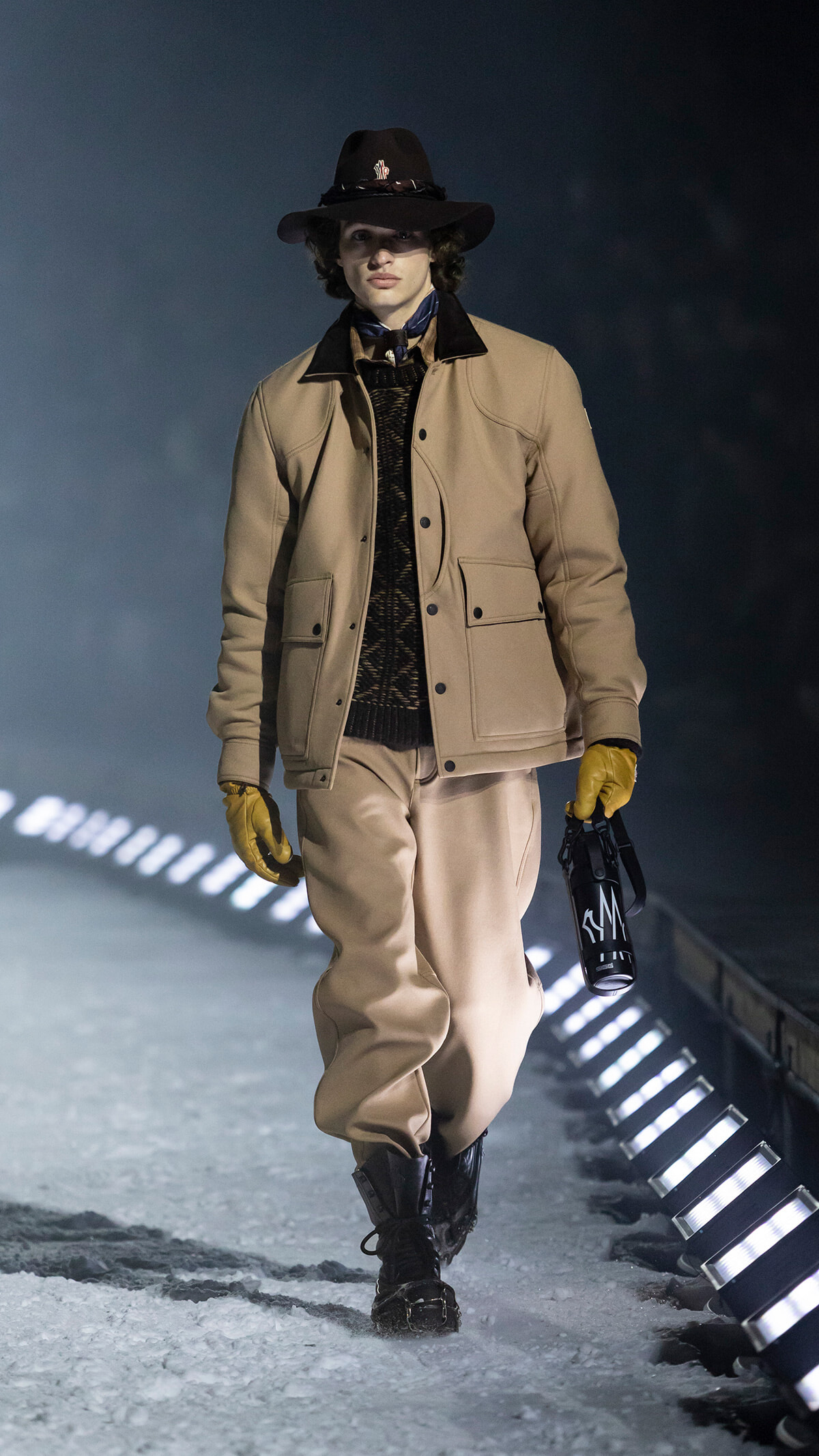 Model walking on a snowy runway in a tan beige jacket, matching pants, yellow gloves, and a black hat, holding a black bottle.