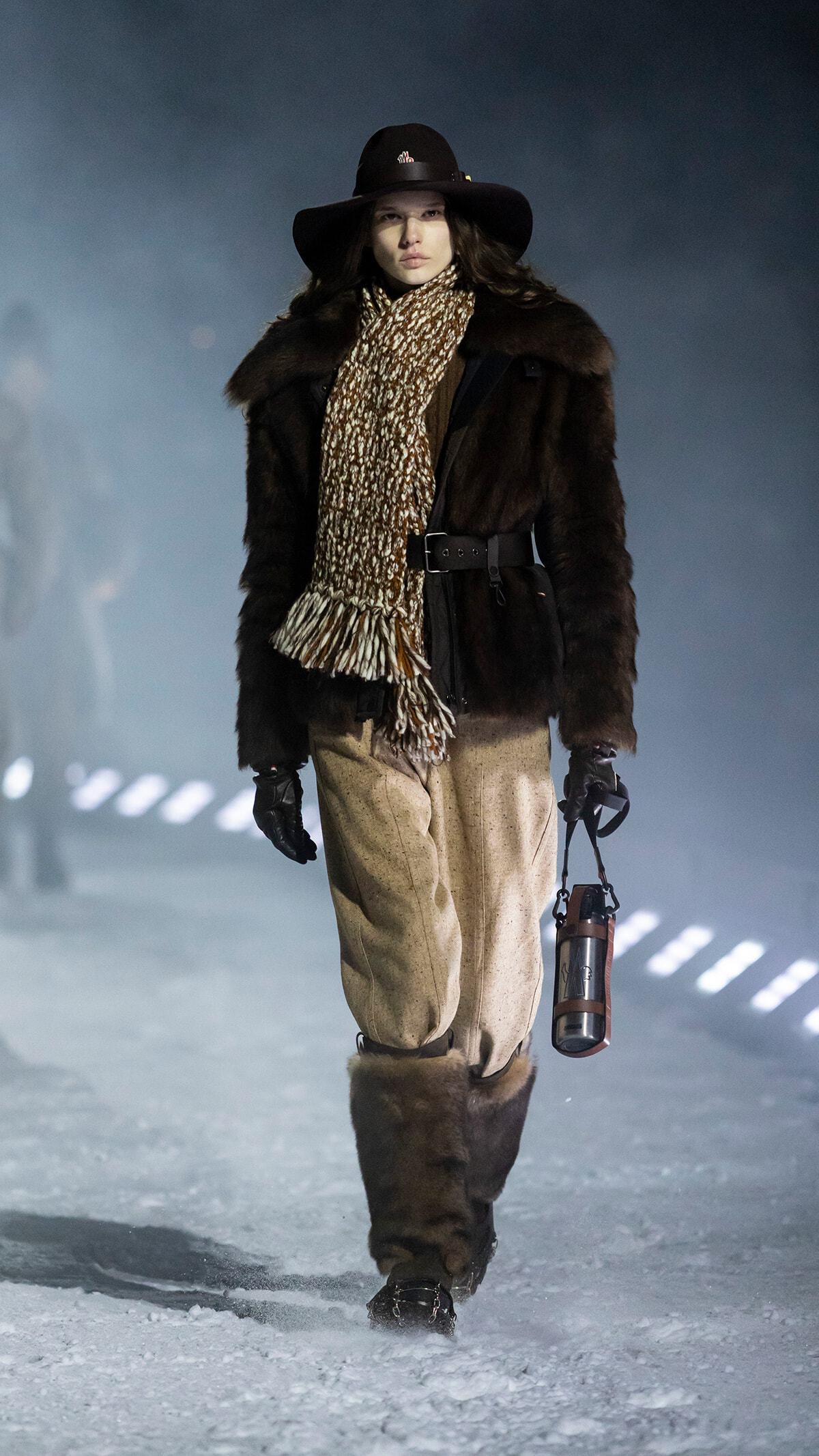 Model walking a snowy runway in a dark fur coat, wide-brim hat, chunky leopard scarf, tan wool pants, and fur-lined boots, carrying a water bottle.