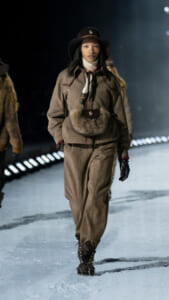 Female model walking a winter runway in a brown wool suit, fur belt pouch, wide-brim hat, and scarf.