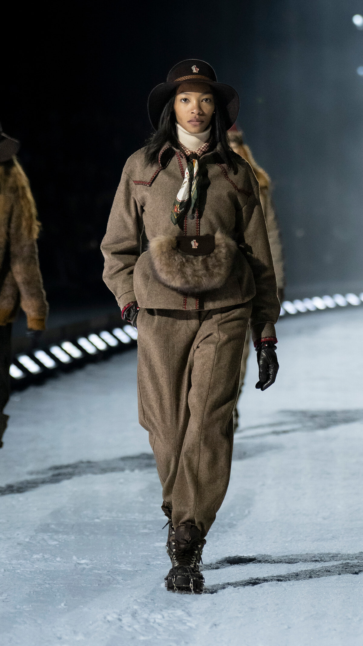 Female model walking a winter runway in a brown wool suit, fur belt pouch, wide-brim hat, and scarf.
