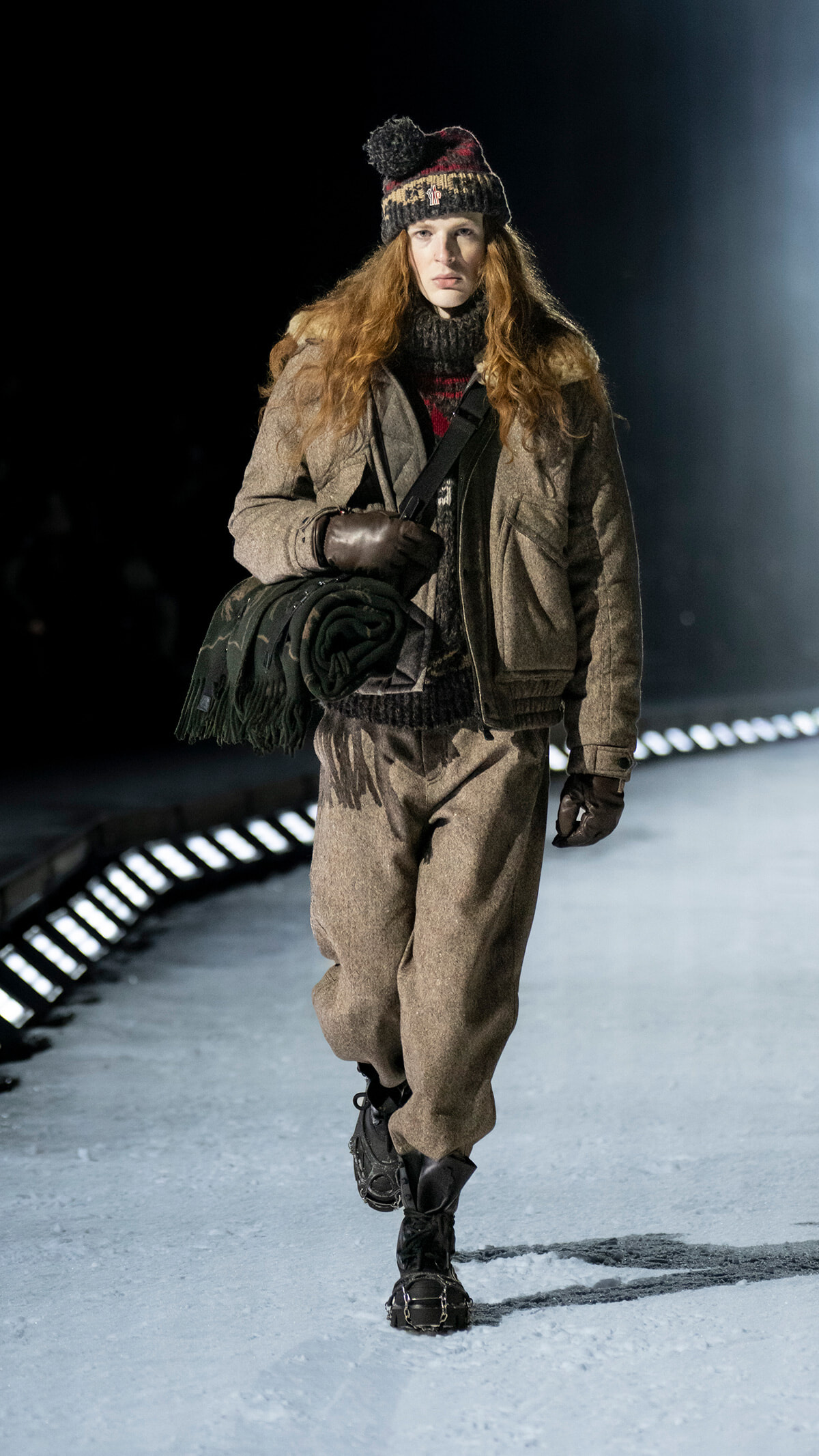 Female model walks a snowy runway in a tan wool jacket, chunky knit hat, brown gloves, with a bag strap and a rolled green blanket over her arm.