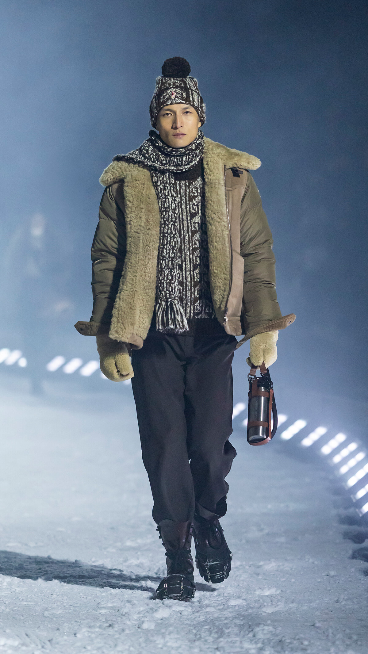 Person walking in heavy winter coat with shearling lining, hat, scarf, and gloves on a snowy path, carrying a thermos.