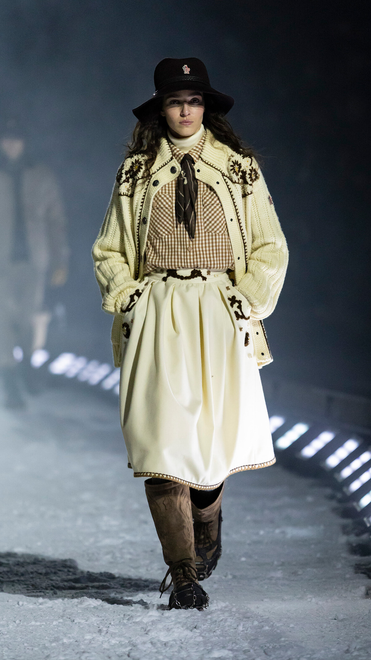 Model walking a snowy runway in a cream cardigan with floral embroidery, a checkered blouse with a tie, and a full cream skirt paired with brown boots and a black hat.
