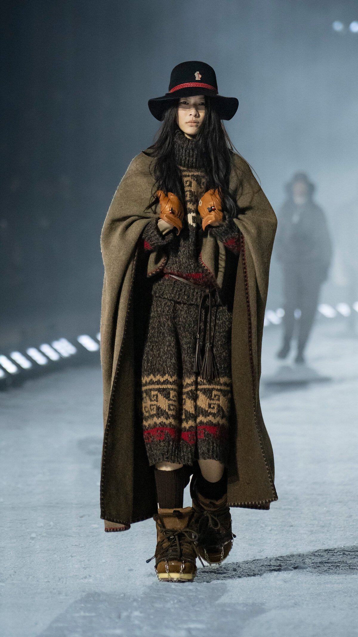 Model walking a runway in a chunky knit sweater with a brown patterned poncho and matching knit pants, topped with a black wide-brim hat and brown gloves.