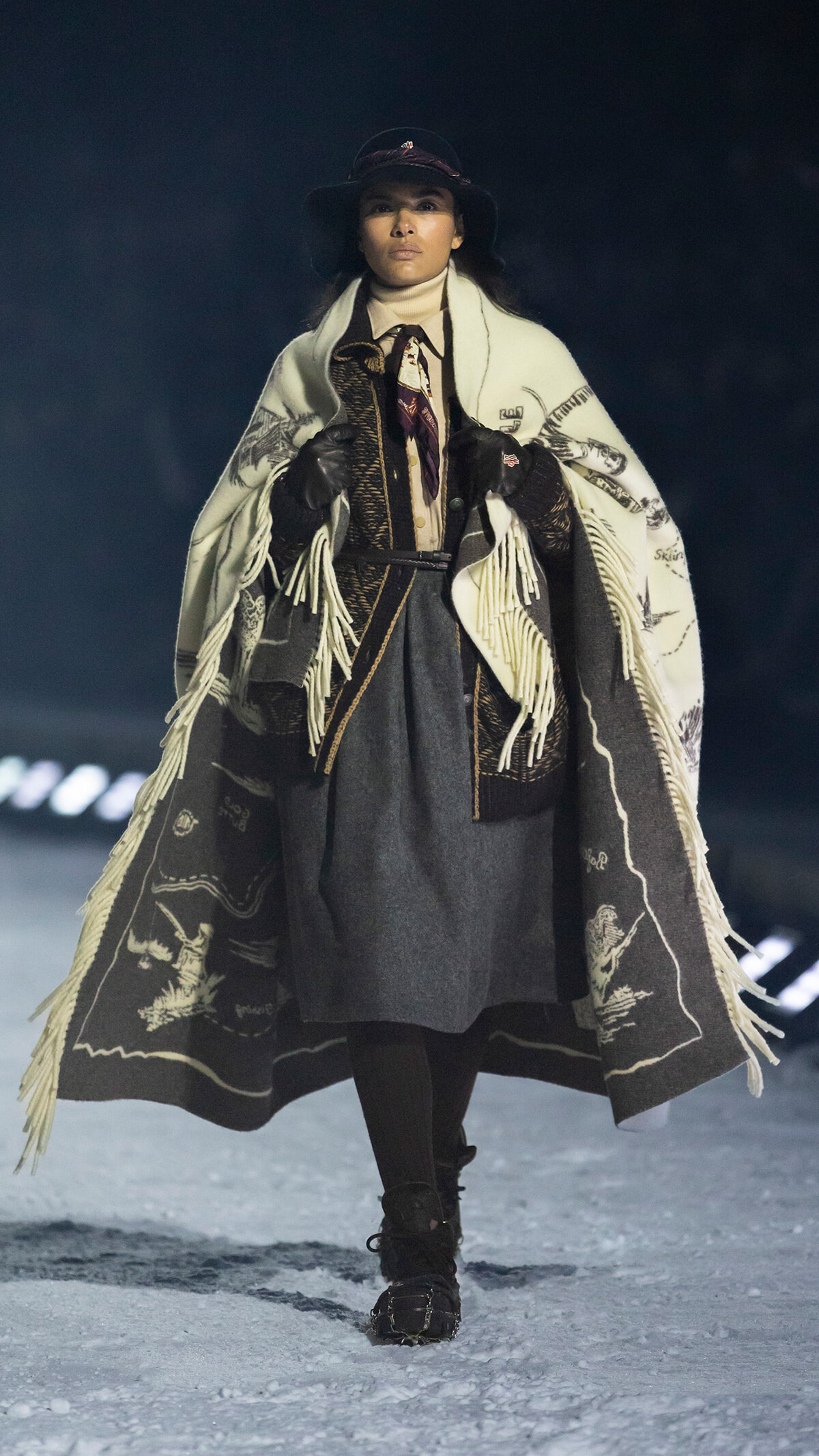 Model walking on a runway in a layered Western-inspired outfit: fringed cream poncho over brown vest and dark skirt, with hat and gloves.