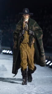 Male model walking a snowy runway in a dark wide-brim hat, green patterned poncho, tan layered outfit, gloves, and sturdy boots.