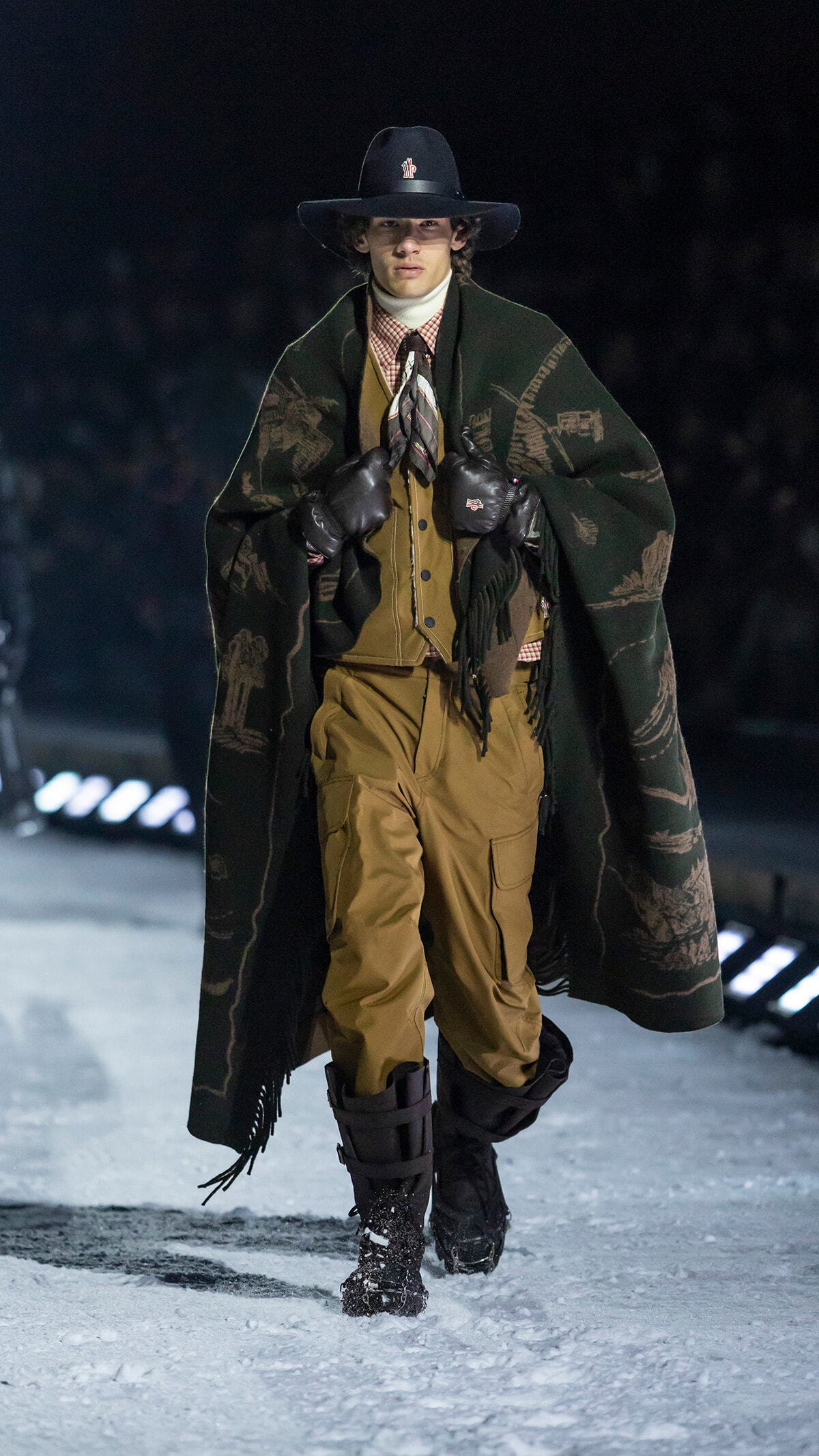 Male model walking a snowy runway in a dark wide-brim hat, green patterned poncho, tan layered outfit, gloves, and sturdy boots.